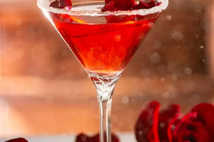 Red cocktail in a sugar-rimmed glass with rose petals and strawberries.