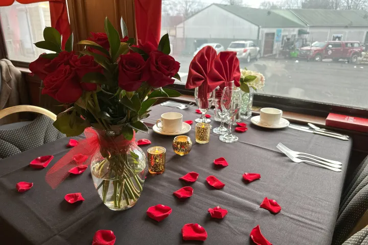 Table set with red roses, candles, and scattered petals near a window.