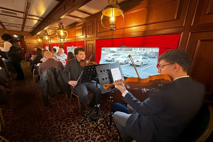 Musicians playing in a cozy dining car with diners seated, red curtains, and parked cars visible outside.