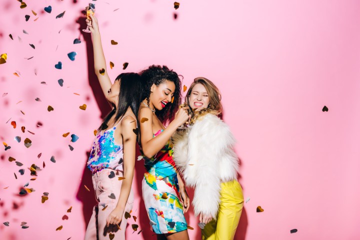 Three women celebrating with confetti and drinks against a pink background.