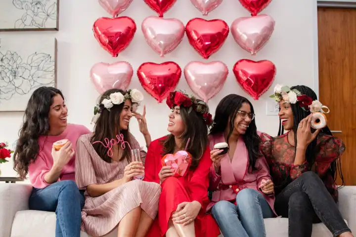 Five women celebrate with heart balloons and food, wearing flower crowns and smiling on a sofa.