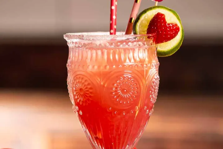 Ornate glass of pink drink with straws, lime and strawberry garnish on pink cloth.