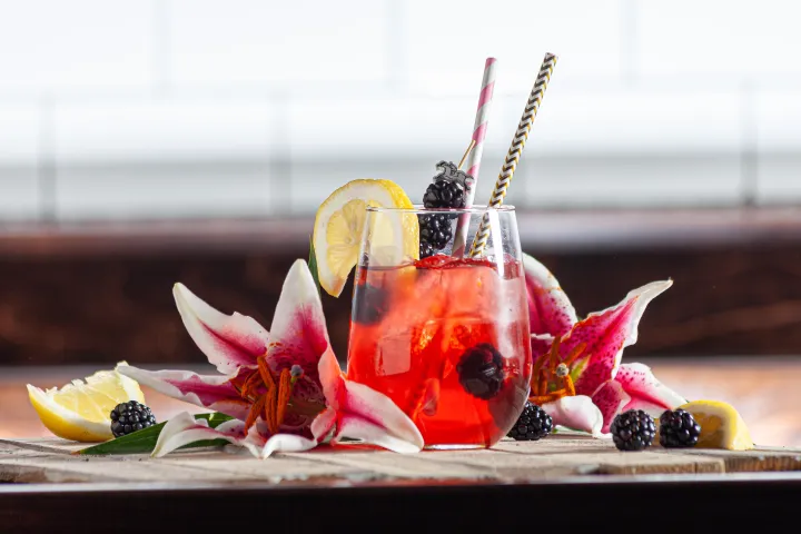 Red drink with lemon slices, blackberries, and pink lilies in a glass with straws on a wooden surface.