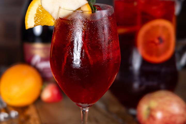 Red cocktail in a glass with orange slice, mint, apple, strawberries, and bottle on a wooden table.