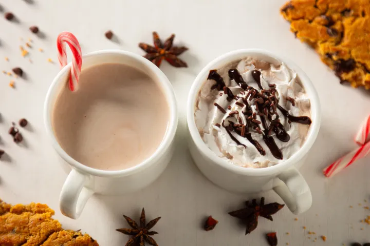 Two mugs of hot chocolate, one with whipped cream and chocolate drizzle, with candy canes and cookies nearby.