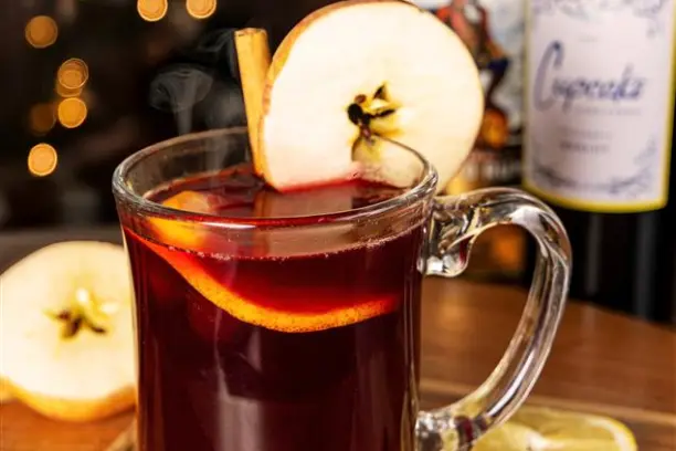 Glass mug of mulled wine with apple slice and cinnamon, bottles in background, on wooden surface.
