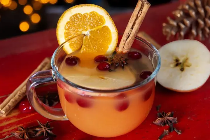 Warm holiday drink in a glass mug with orange slice, cranberries, and cinnamon on a red table.