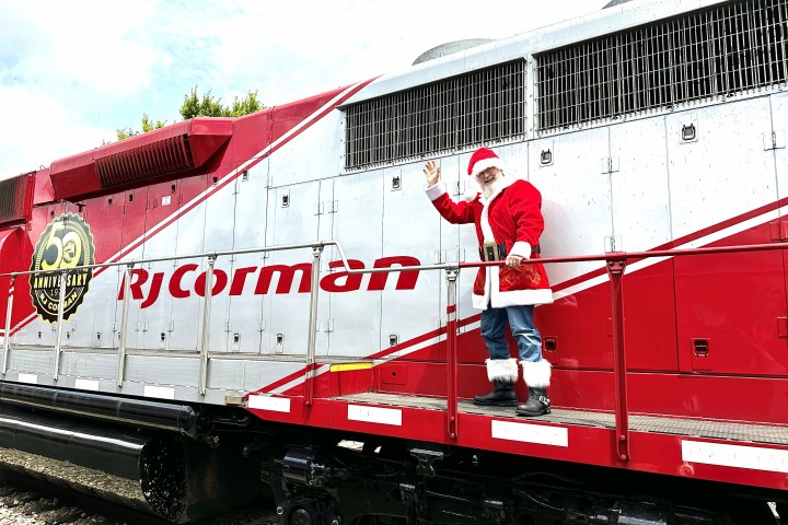 Person in Santa outfit waves from a red train labeled 'RJ Corman'.